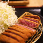 What to Eat in Miyazaki: 6 Must-Try Dishes A plate with sliced breaded beef cutlet, cooked rare—a Miyazaki must-try dish—beside a generous serving of finely shredded cabbage on a wire rack, with dipping sauce in the background. Wondering what to eat? Start here.