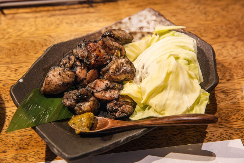 A black plate with grilled chicken pieces, raw cabbage leaves, a green condiment, and a wooden spoon set on a wooden table—one of Miyazaki’s must-try dishes and a perfect answer to what to eat in this region.