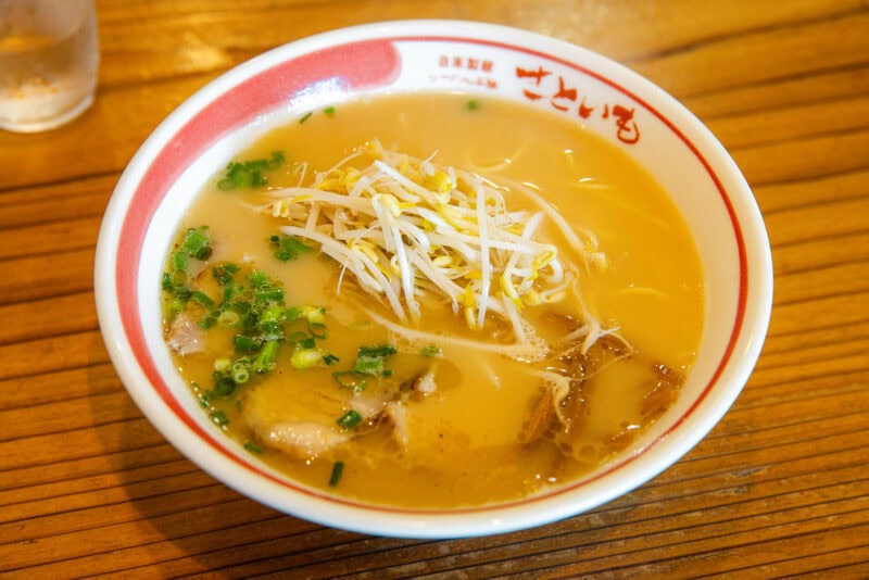 A bowl of ramen with creamy broth, topped with sliced green onions, bean sprouts, and pieces of pork, served in a white bowl with red trim on a wooden table—one of Miyazaki’s must-try dishes for anyone wondering what to eat.