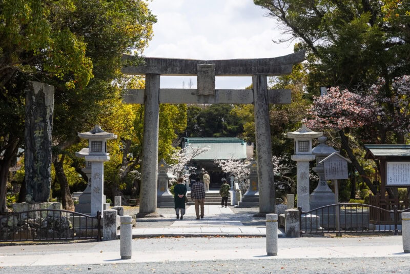 A stone torii gate leads to Munakata Shrine, one of Japan's oldest shrines, surrounded by trees and stone lanterns. People walk along the pathway under blooming cherry blossoms on a sunny day.