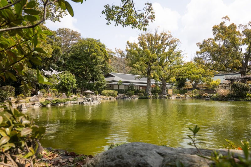 A serene Japanese garden with a large pond, lush trees, rocks, and a traditional wooden building—reminiscent of Japan's oldest shrines—rests peacefully under a partly cloudy sky.