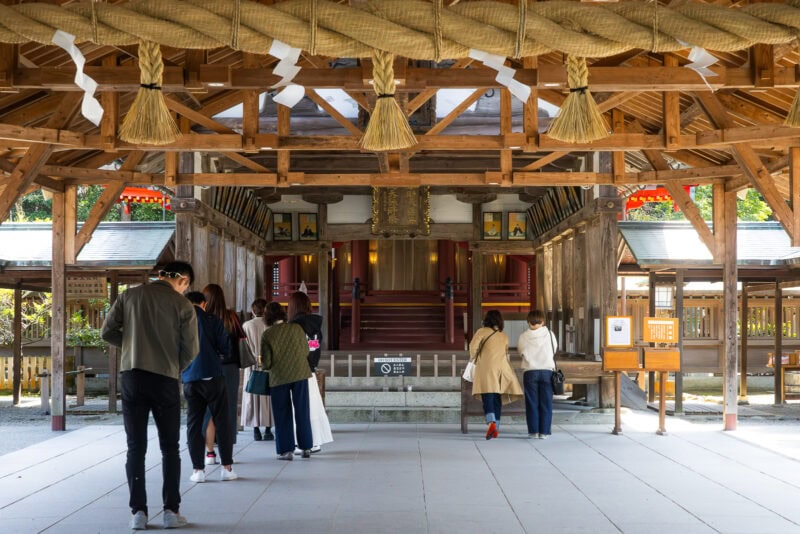 People stand and bow at Munakata Shrine, Japan's oldest shrine, surrounded by traditional architecture, straw decorations, and hanging ornaments, engaging in prayer or respectful observation. A guide may share insights into these sacred rituals.