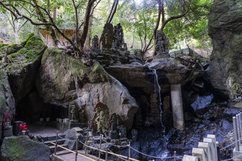 Stone statues and carvings are arranged on rocky cliffs near a small waterfall in a forested area at Nanzoin in Fukuoka, with the famous Reclining Buddha nearby, a wooden bridge in the foreground, and lush green trees overhead.