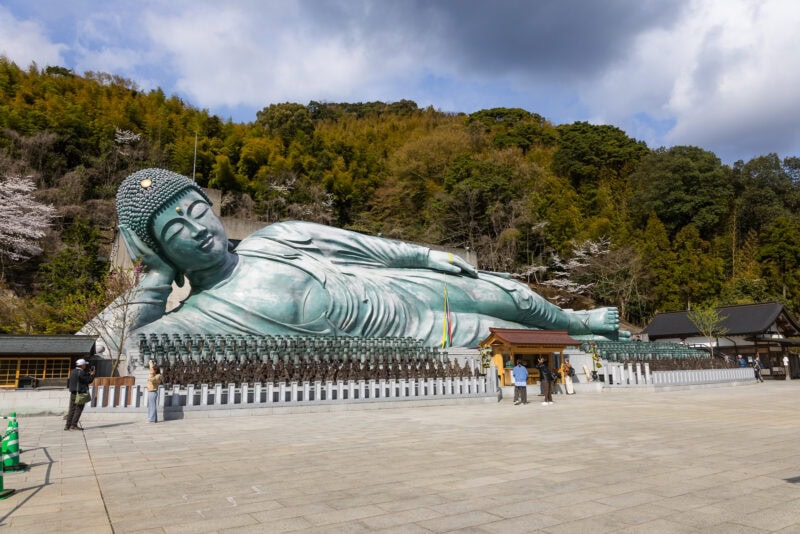 The Nanzoin Reclining Buddha, one of Fukuoka’s iconic landmarks, lies outdoors surrounded by trees, with small statues lined up in front. Visitors stroll nearby, taking photos under a partly cloudy sky.