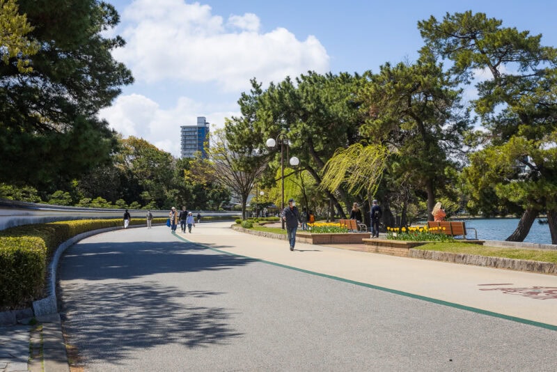 A paved park path in Ohori Park, Fukuoka, lined with trees and flower beds, curves alongside the water. People walk, sit on benches, and enjoy a sunny day in this urban oasis, with a building rising in the background.