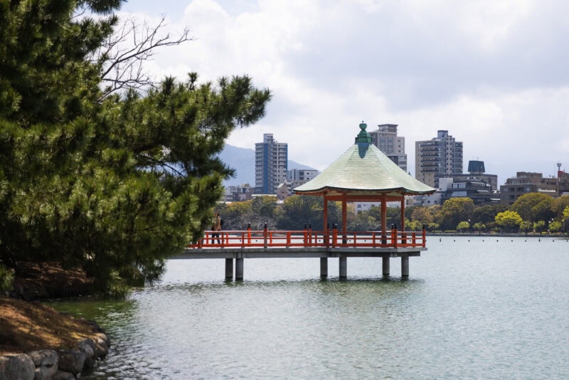 A traditional Japanese pavilion with a green roof stands over a calm lake in Ohori Park, Fukuokaāan urban oasis surrounded by trees, with modern city buildings visible in the background under a partly cloudy sky.