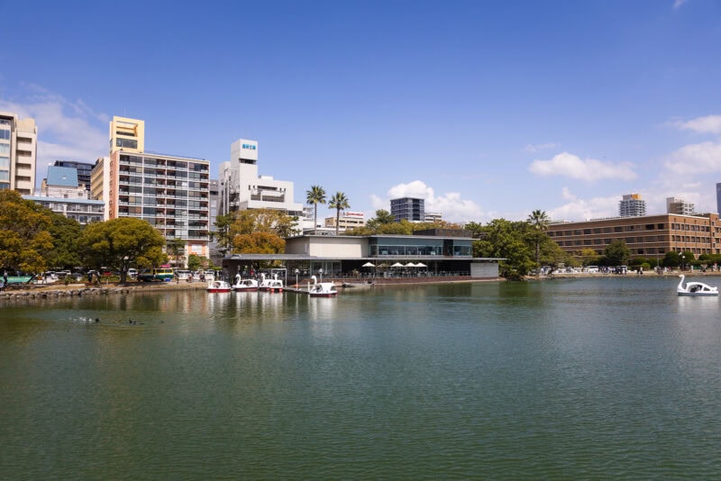 A calm urban oasis, Ohori Park in Fukuoka features swan boats docked along the shore, surrounded by modern buildings, trees, and a blue sky with scattered clouds. People can be seen relaxing near the waterfront.