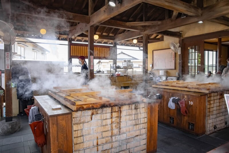 A traditional Japanese kitchen in Oita with steaming wooden-lidded pots on brick stoves, wooden beams overhead, and people working in the background. Fans and a menu board highlight some must-try dishes for those curious about what to eat.