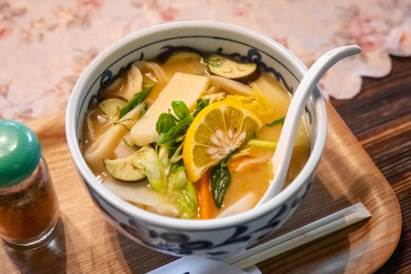 A bowl of noodle soup topped with sliced vegetables and a lemon wedge, with a ceramic spoon resting inside. Chopsticks and a spice shaker sit on the wooden tray—one of Oita’s must-try dishes if you’re wondering what to eat.