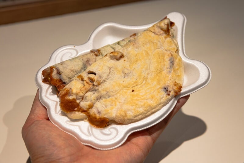 A hand holding a white, shell-shaped disposable plate with two folded, golden-brown savory pancakes, likely filled with vegetables and meat—an appetizing highlight from any Osaka food guide.