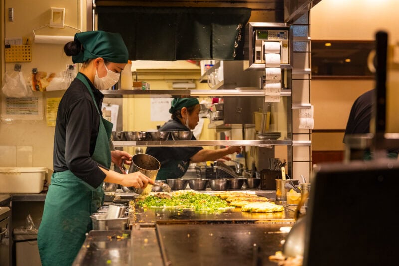 A chef in a green apron and headscarf prepares food on a large grill in a restaurant kitchen, adding ingredients to savory pancakes with various toppings—a scene straight from an Osaka food guide. The chef’s reflection is visible in the mirror behind.