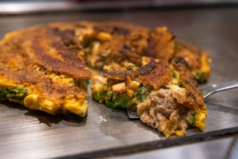 A close-up of a savory pancake, crispy on the outside, with a fork lifting a bite showing meat, green onions, and other fillings—an irresistible scene straight from any Osaka food guide, resting on a hot metal surface.