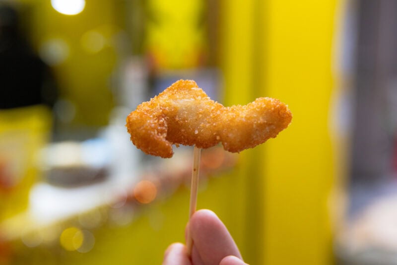 A hand holds a skewer with a golden, breaded, and fried food shaped like a fish, against a blurred, bright yellow background—perfect for any Osaka food guide.