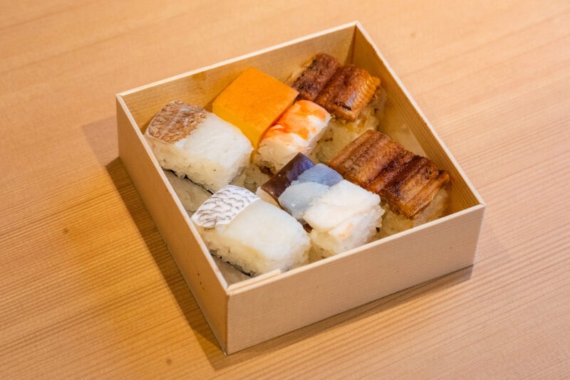 A wooden box containing assorted pressed sushi pieces, including fish and egg toppings, neatly arranged on a light wooden table—a delicious highlight from any Osaka food guide.
