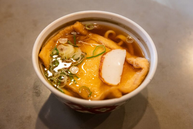 A bowl of udon soup with a slice of fish cake, chopped green onions, and fried tofu in clear broth, served in a patterned cup on a gray surface—a must-try from any Osaka food guide.
