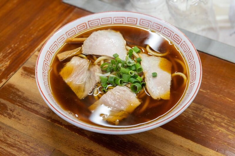 A bowl of ramen with dark soy-based broth, topped with slices of pork, chopped green onions, and bamboo shoots—perfect for any Osaka food guide—served on a wooden table.
