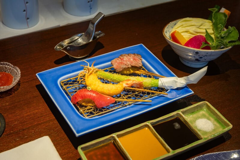 A blue plate holds assorted skewered tempura—shrimp, fish, and a green vegetable—accompanied by sauces, fresh vegetables, and a dipping sauce on a wooden table, perfect for any Osaka food guide.