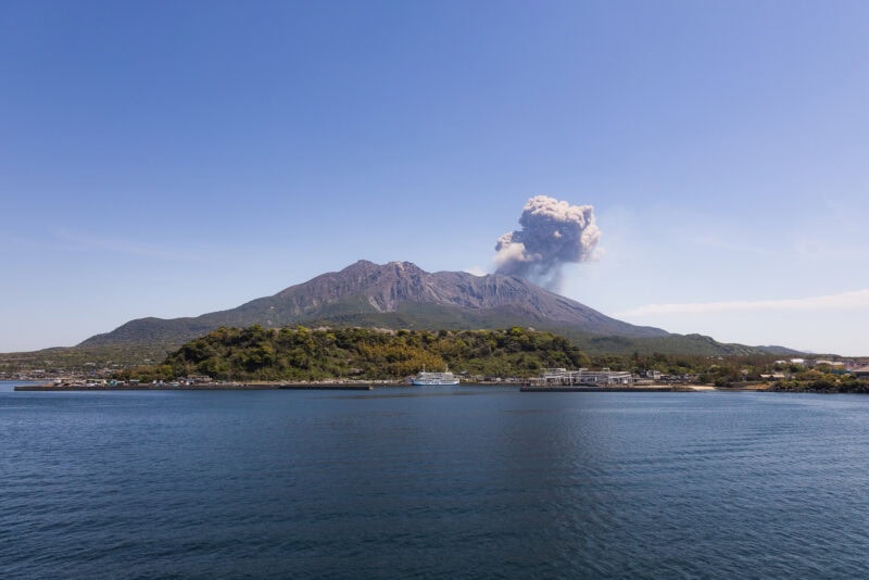 A large volcano, Sakurajima, rises behind the coastal town of Kagoshima, emitting a thick plume of smoke into the clear blue sky, with calm blue water in the foreground&mdash;a powerful symbol of the region's natural beauty.