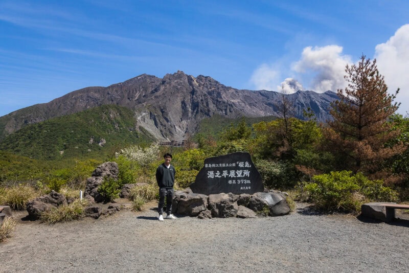A person stands in front of a large black stone marker with Japanese text, surrounded by greenery, with Sakurajima volcano smoking in the background beneath the clear blue sky of Kagoshima.