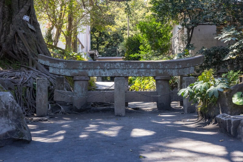 A low, weathered stone torii gate blocks a shaded dirt path surrounded by trees and plants in a peaceful outdoor setting near Sakurajima, Kagoshima. Sunlight filters through the leaves, creating dappled shadows on the ground.