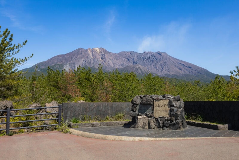 A stone monument stands at a paved lookout in Kagoshima, surrounded by green trees with the majestic Sakurajima volcano rising in the background under a clear blue sky.