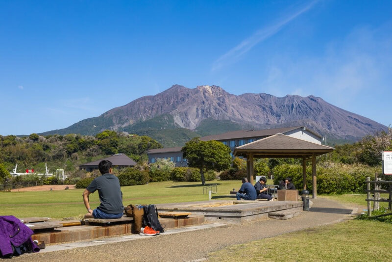 People sit and relax at an outdoor footbath area in a park, enjoying a scenic view of Sakurajima and the clear blue sky. A gazebo and buildings dot the lush Kagoshima landscape, creating a peaceful atmosphere amid nature.