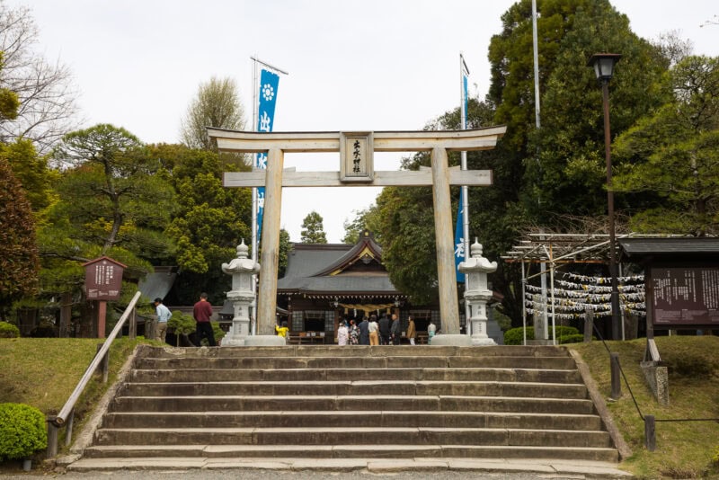 A traditional Japanese torii gate stands at the entrance to a Shinto shrine in Kumamoto, with stone lanterns on either side and steps leading up. People gather near the shrine amidst lush green trees, reminiscent of the tranquil beauty of Suizenji Jojuen historic garden.