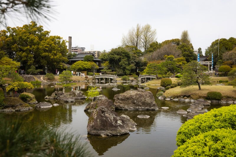 A serene Japanese garden, Suizenji Jojuen in Kumamoto, features a pond, large rocks, manicured bushes, and an arched stone bridge by a traditional-style building&mdash;an iconic historic garden surrounded by lush green trees.