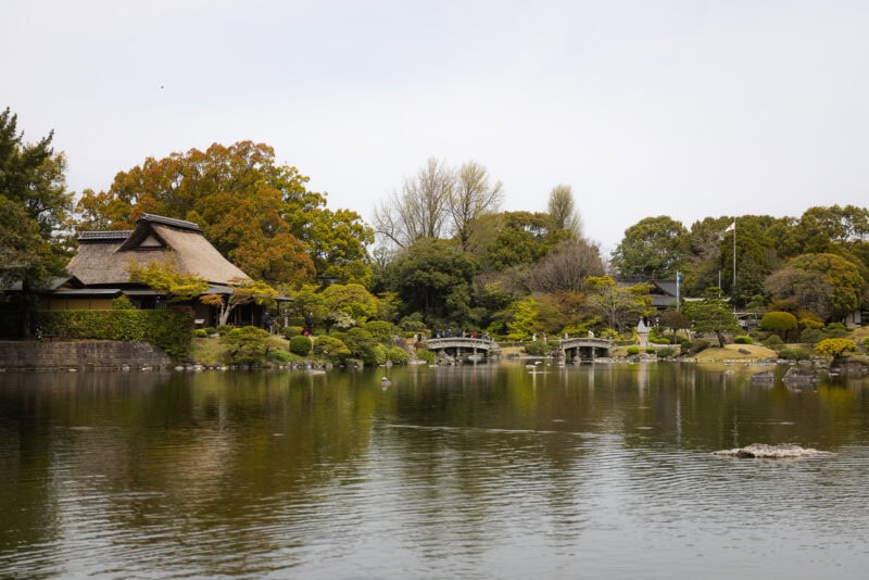 A serene Japanese garden, Suizenji Jojuen in Kumamoto, features a large pond, arched stone bridges, lush green trees, and a traditional thatched-roof building along the water’s edge under a cloudy sky.