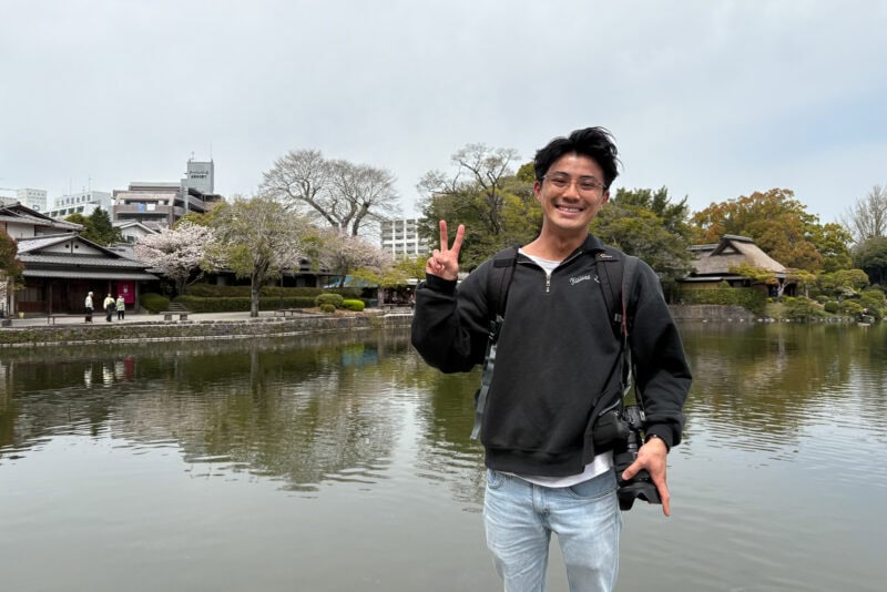 A man with glasses and a camera around his neck smiles and makes a peace sign while standing in front of the historic garden Suizenji Jojuen in Kumamoto, with traditional Japanese buildings and cherry blossom trees in the background.