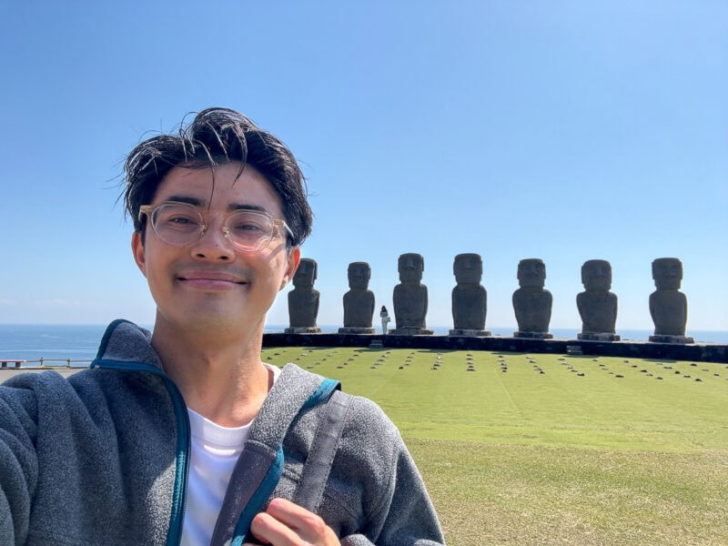 A person with glasses smiles for a selfie in front of six large Moai statues at Sun Messe Nichinan, Japan's Easter Island, on a grassy field with a clear blue sky and ocean in the background.
