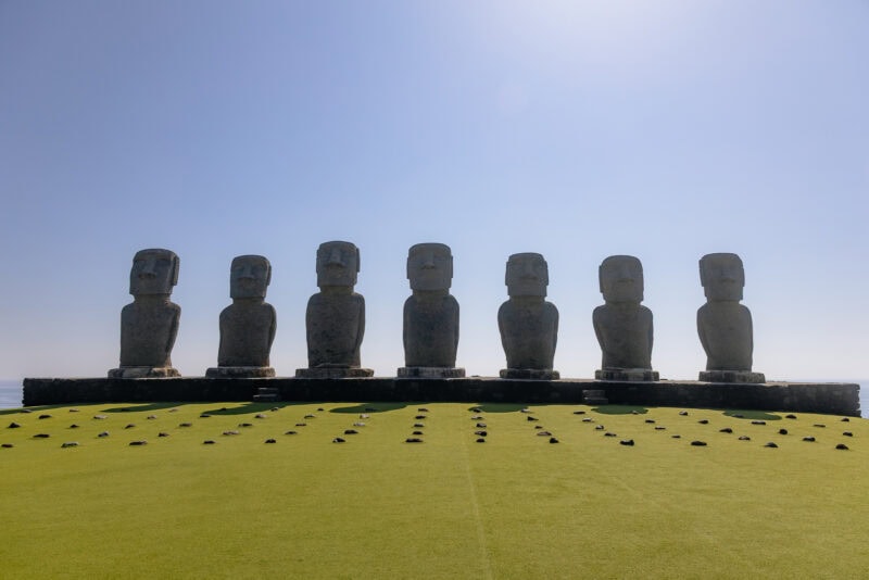 Seven large stone Moai statues stand in a row on a stone platform at Sun Messe Nichinan, Japan's Easter Island, with a clear blue sky and green grass in the foreground. The statues face forward in an open, sunny landscape.