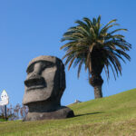 Sun Messe Nichinan: Japan's Easter Island A large stone Moai statue sits on a grassy slope near a tall palm tree under a clear blue sky at Sun Messe Nichinan, Japan's Easter Island, with a cartoonish sign visible in the background.