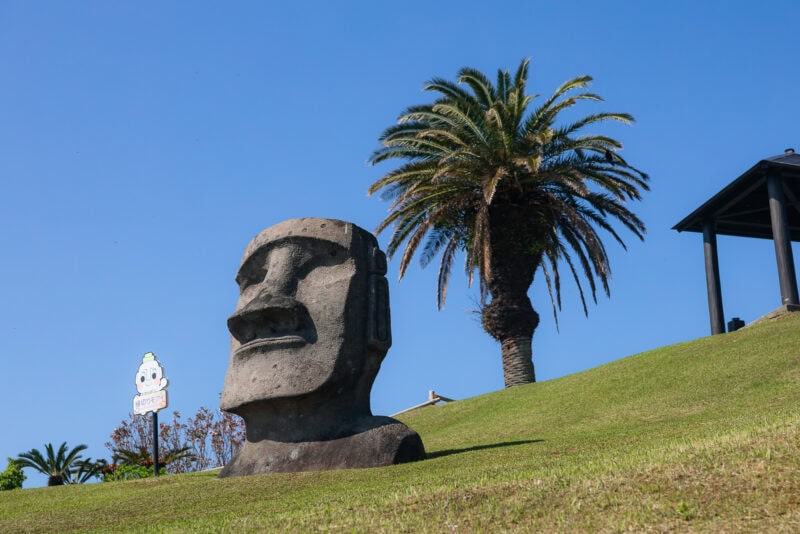 A large stone Moai statue sits on a grassy slope near a tall palm tree under a clear blue sky at Sun Messe Nichinan, Japan's Easter Island, with a cartoonish sign visible in the background.