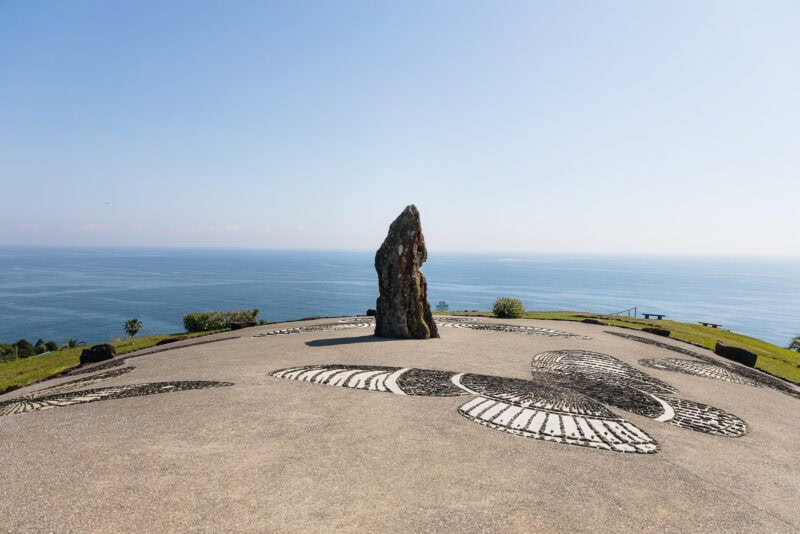 A tall stone stands upright on a circular concrete platform at Sun Messe Nichinan, known as Japan's Easter Island, decorated with large bird designs and overlooking a calm blue ocean under a clear sky. Green grass surrounds the platform.