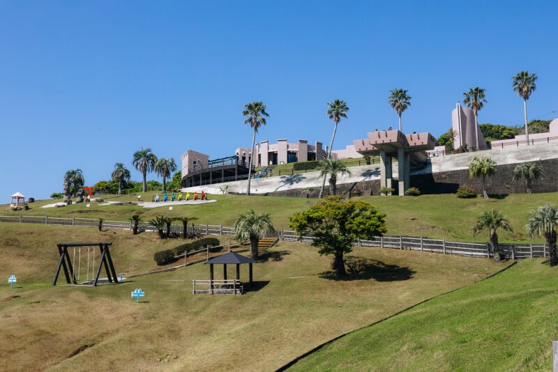 A grassy hillside park with palm trees, swings, shaded seating areas, and modern buildings at the top—Sun Messe Nichinan, known as Japan's Easter Island, sits beneath a clear blue sky.
