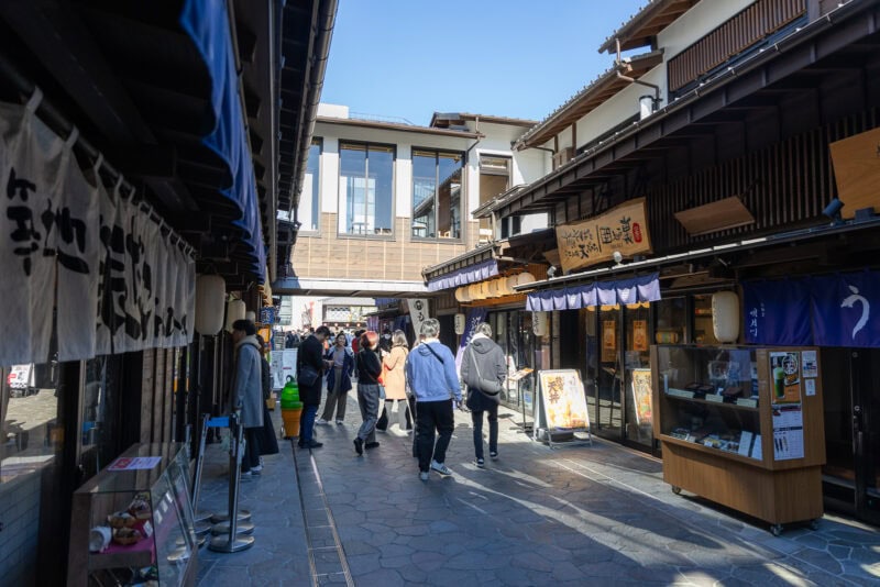 A group of people walk along a narrow, stone-paved street lined with traditional Japanese wooden shops and restaurants at Toyosu Senkyaku Banrai, with signs and menus displayed outside under a clear blue sky.