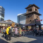 Toyosu Senkyaku Banrai: Tokyo's Edo Food Village Guide People walk and sit near a tall wooden tower and outdoor market stalls at Toyosu Senkyaku Banrai on a sunny day; modern buildings and red lanterns are in the background.