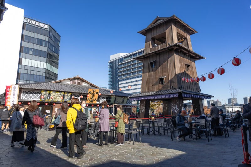 People walk and sit near a tall wooden tower and outdoor market stalls at Toyosu Senkyaku Banrai on a sunny day; modern buildings and red lanterns are in the background.