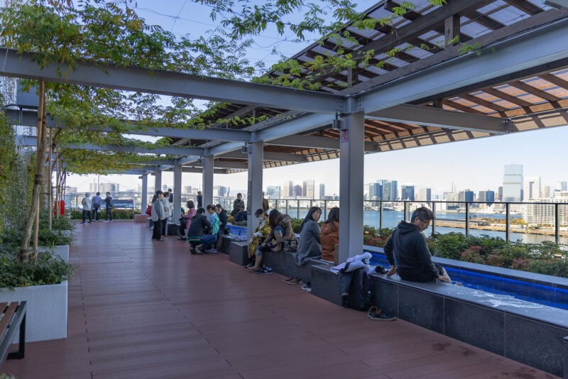 People sit and relax along the rooftop terrace at Toyosu Senkyaku Banrai, featuring a long water feature and views of the city skyline. The wooden floor and pergolas with leafy vines provide inviting shade under a clear sky.