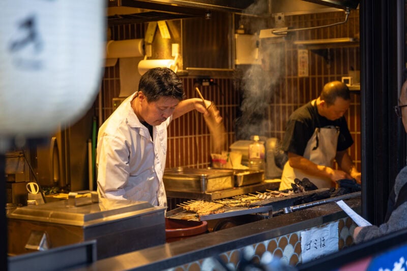 At Toyosu Senkyaku Banrai, a chef grills skewers of meat over an open flame at a bustling food stall, while another person works in the background. Smoke rises from the grill as a customer waits at the counter in the foreground.