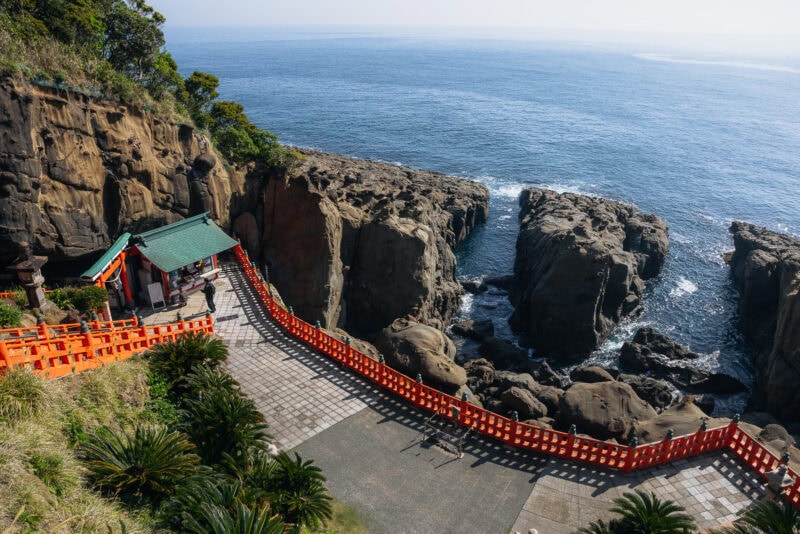 The Udo Shrine, a scenic seaside shrine in Miyazaki, features a green roof perched atop rocky cliffs, framed by orange railings and lush greenery, offering breathtaking ocean views under a clear sky.