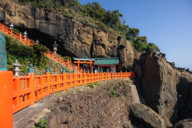 A vibrant orange railing lines a stone pathway leading to Udo Shrine, a seaside shrine built into a rocky cliffside in Miyazaki, surrounded by lush greenery under a clear blue sky.