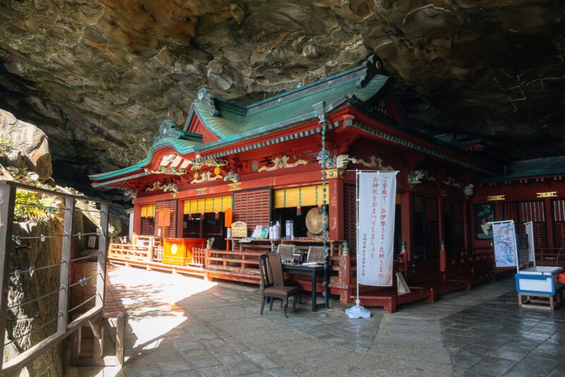 A traditional Japanese shrine with green curved roofs and red wooden walls is built beneath a rocky overhang. Sunlight filters in, illuminating the entrance and stone floor of Udo Shrine, a famous seaside shrine in Miyazaki.