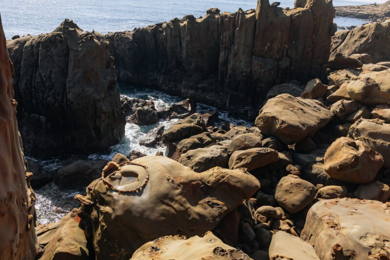 Rocky coastal cliffs with large, weathered boulders and ocean waves crashing below. In the foreground, a circular structure with sharp teeth-like objects is visible on a flat rock near the famed Udo Shrine, a unique seaside shrine in Miyazaki.