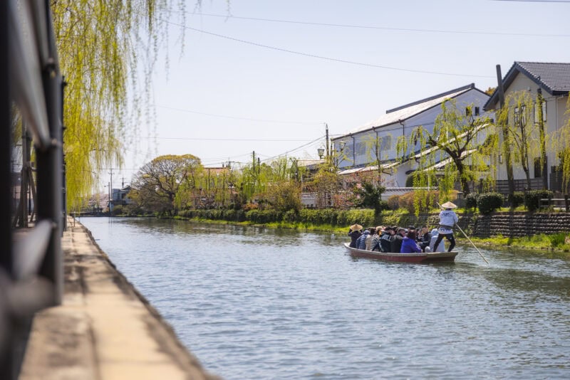A group of people ride in a small wooden boat steered by a guide on the calm Yanagawa Canals, known as the "Venice of Kyushu," lined with green willow trees and traditional houses on a sunny day.