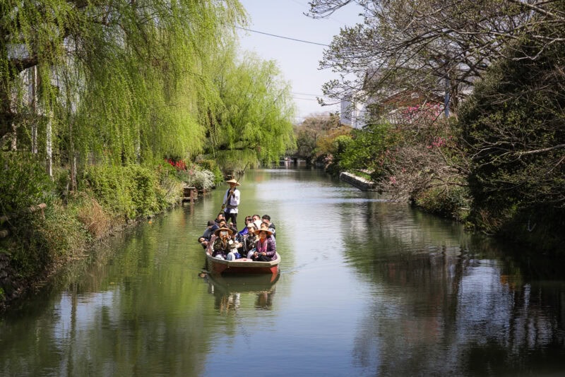 A small group of people sits in a boat on the Yanagawa Canals, known as the "Venice of Kyushu," gliding along a calm, narrow waterway lined with lush green trees and foliage under a sunny sky.