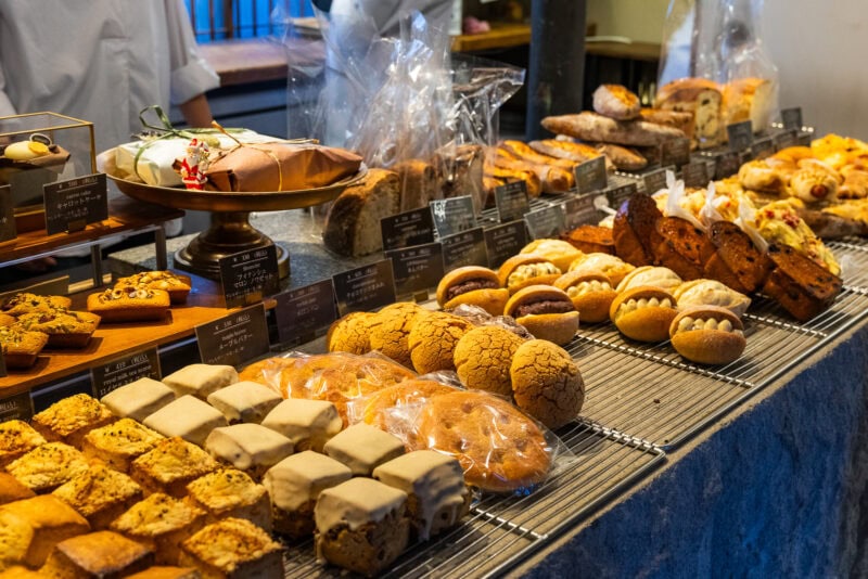 A bakery display in Yanaka Ginza features various breads, pastries, and cakes arranged on metal racks and trays, each labeled with signs. Wrapped loaves, cookies, and tarts are visible, with bakers standing behind the counter.