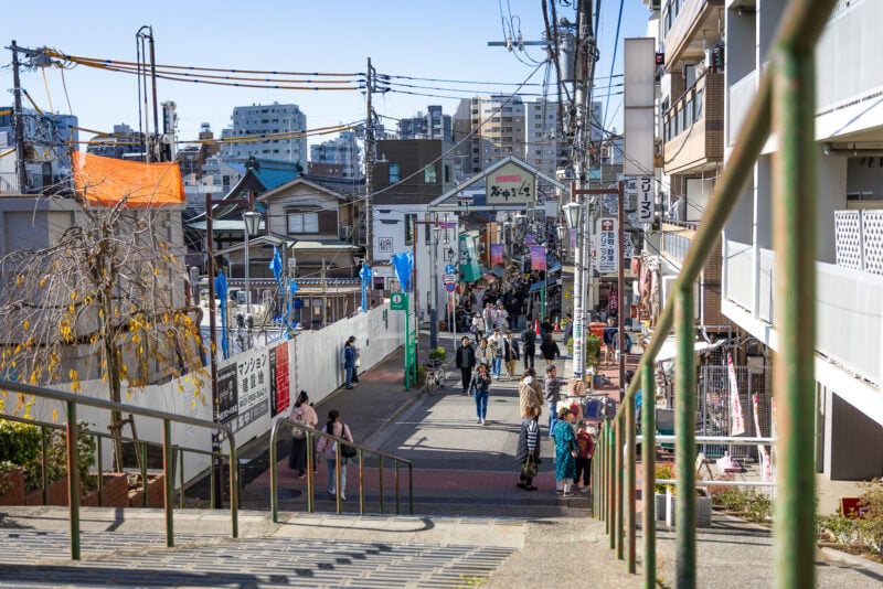 A bustling street in Yanaka Ginza, viewed from the top of a staircase. People walk along the shops and buildings under clear blue skies, with signs in Japanese and overhead power lines completing the lively scene.