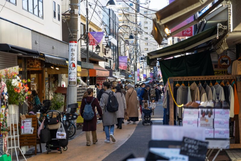A bustling outdoor shopping street, Yanaka Ginza in Japan, with people walking—some with children. Shops line both sides, selling clothes and goods. Signs with Japanese text hang above, and apartment buildings are visible in the background.